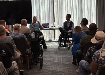 Speaker with a microphone and a fellow panelist in a wheelchair address a seated audience.
Community event in a bright meeting room with attendees, including wheelchair users.